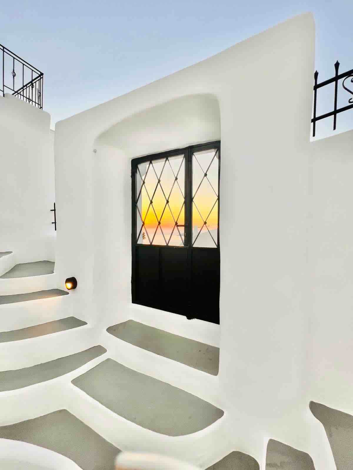 A doorway framed by whitewashed walls leads to a striking sunset view. The staircase features curved steps, blending seamlessly with the surrounding architecture. The glass-paneled door showcases the vibrant colors of the horizon, creating a serene start to evening reflections.