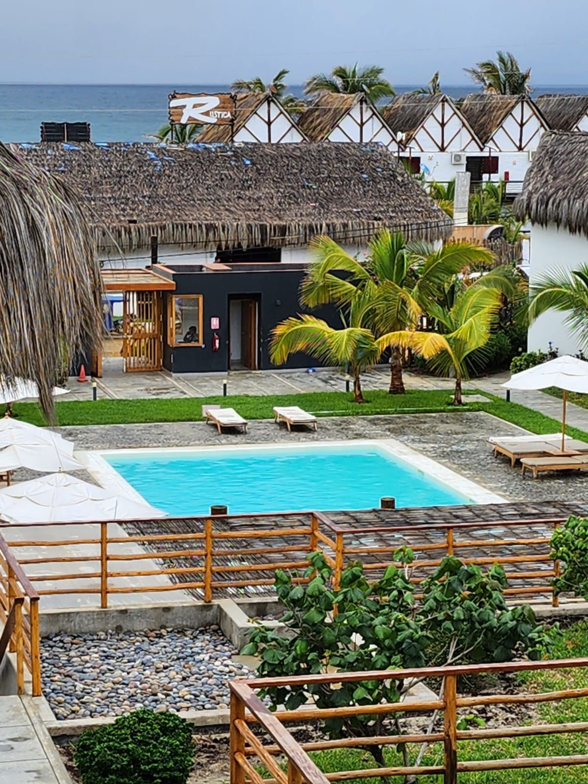A serene outdoor area features a refreshing pool surrounded by lounge chairs and palm trees. The landscape includes well-maintained greenery and a sandy path leading to the beach, with thatched-roof structures in the background.