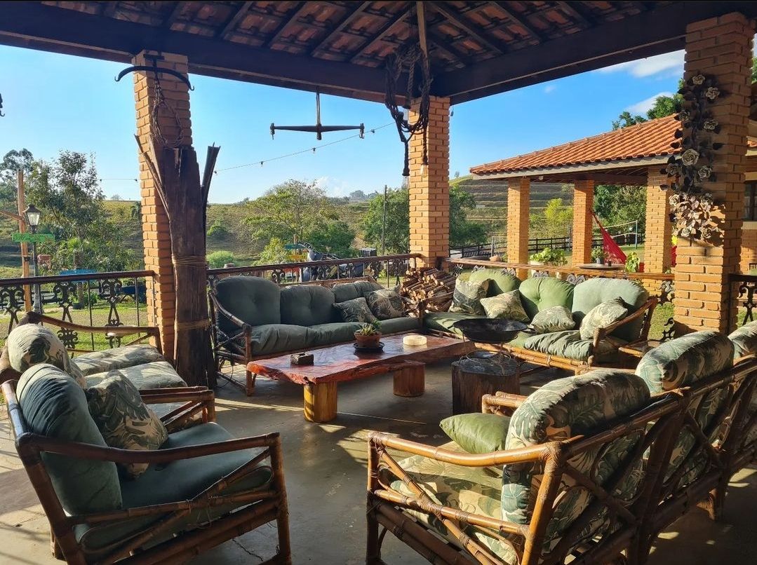 A spacious outdoor lounge area is adorned with comfortable seating arranged around a low wooden table. The setting is framed by brick columns and offers views of the surrounding lush landscape and blue sky.