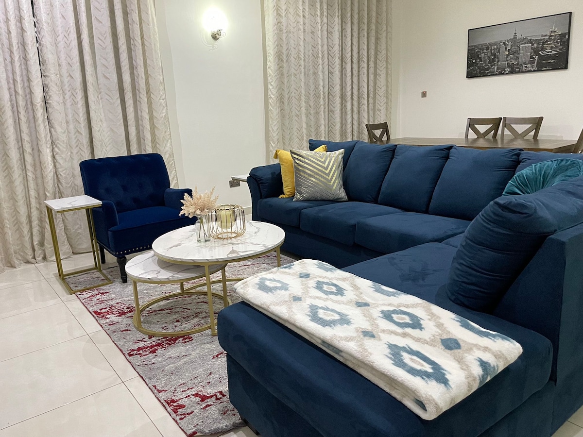 A cozy living area is presented with a navy blue sectional sofa adorned with various cushions. A round coffee table sits at its center, accompanied by a small side table. Light-colored curtains frame the large windows, and a soft area rug adds warmth to the space.