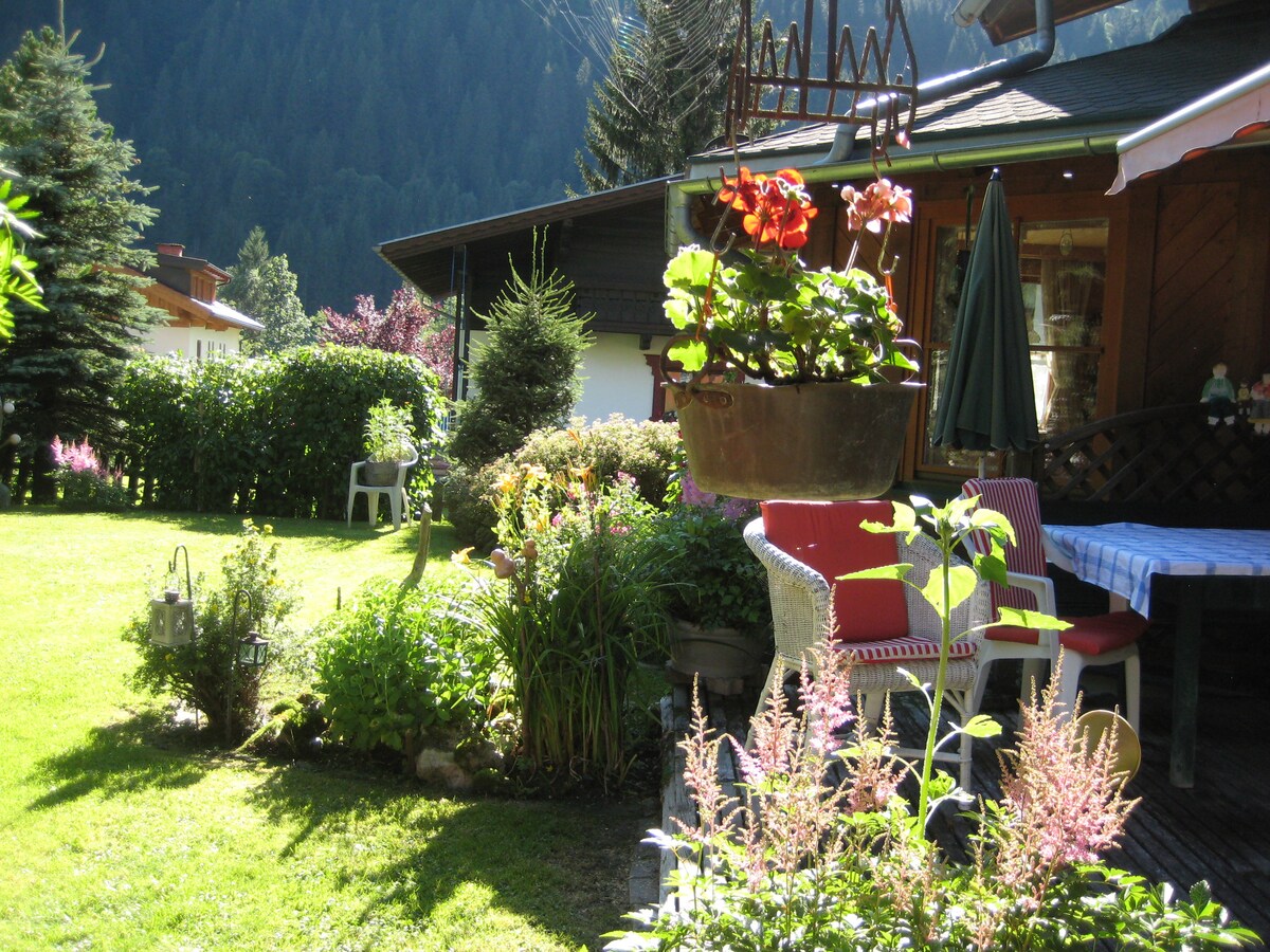 A vibrant garden area features a variety of flowers and greenery, with a cozy seating arrangement including two red chairs and a small table. A hanging flower pot adds color above the setting, surrounded by well-maintained lawns and mountain views in the background.