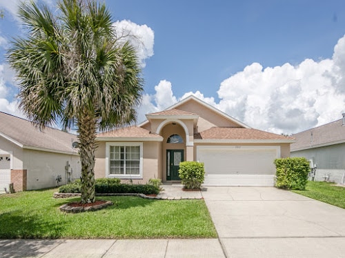A charming single-story villa is presented with a well-maintained front yard featuring a palm tree and manicured shrubs. A paved driveway leads to a two-car garage, while large windows invite natural light into the home. The blue sky contrasts with the architectural details.