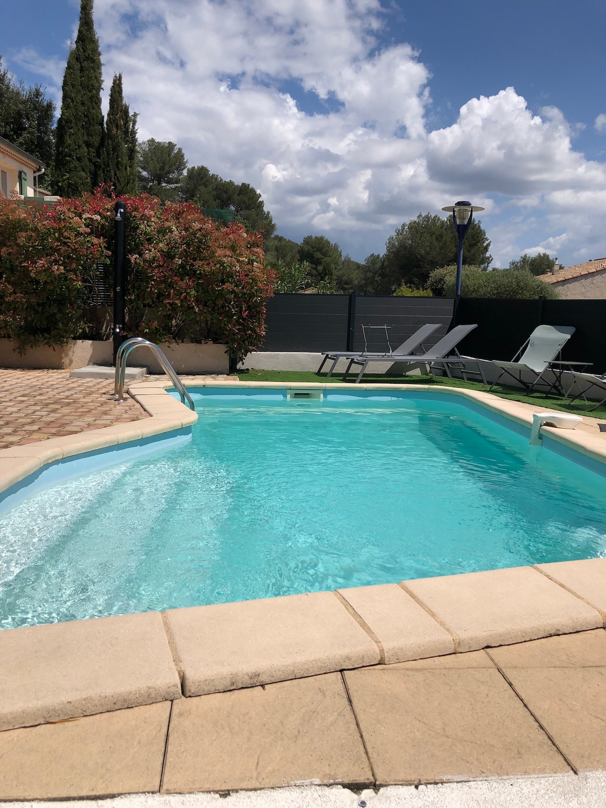 A private swimming pool is surrounded by a well-maintained garden featuring flowering shrubs and a bordered fence. Poolside lounge chairs are positioned nearby for relaxation. The clear blue water invites cooling off on warm days, while a pathway leads to the entrance of the property.