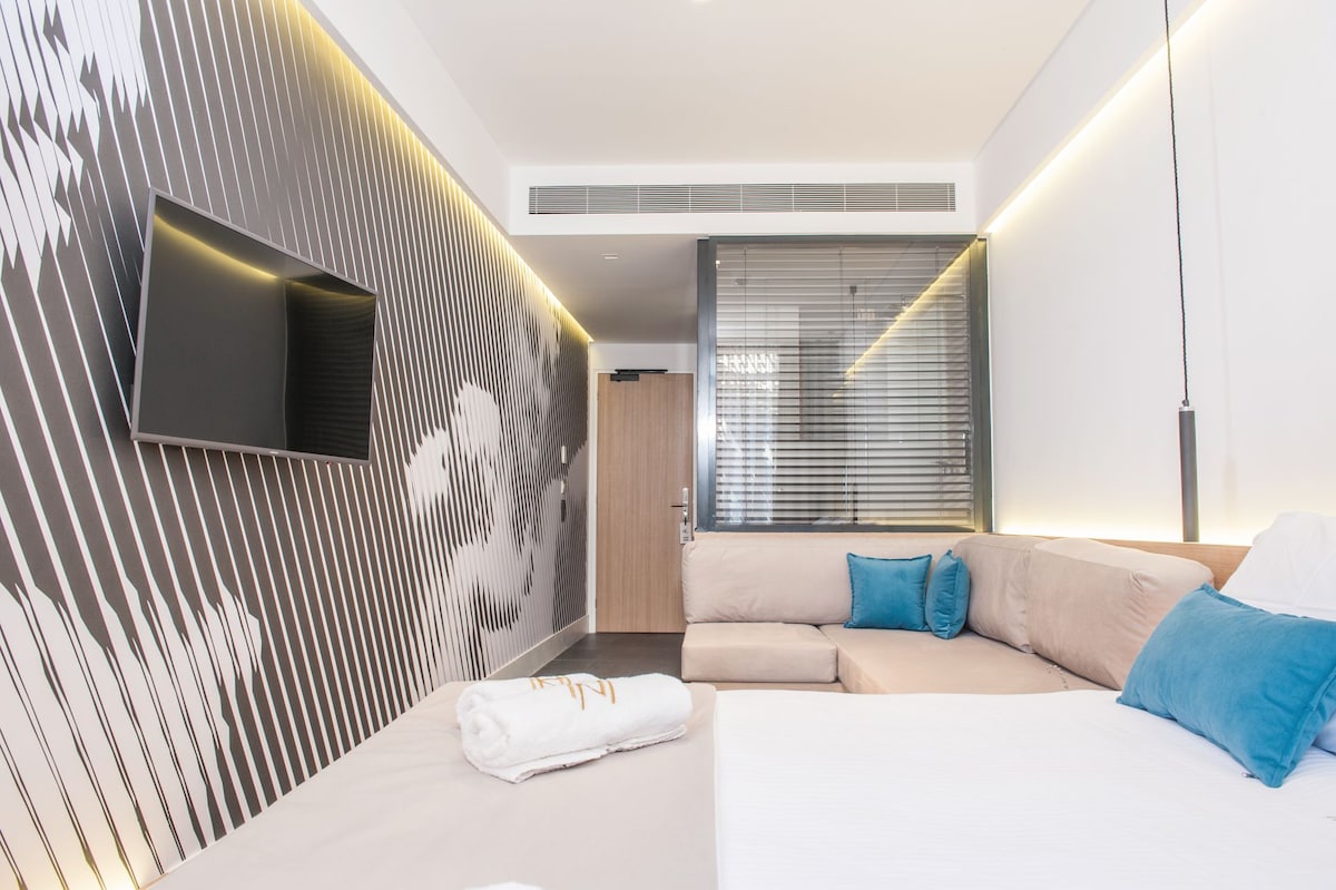 A contemporary room features a large sectional sofa adorned with blue cushions, alongside a spacious bed with neatly arranged towels. A flat-screen TV is mounted on a striped accent wall, while glass partitions provide a modern touch and separation from the entrance.