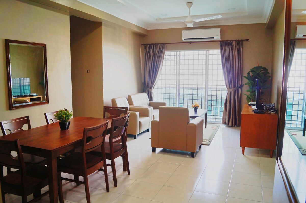 The living area features a comfortable seating arrangement with light-colored sofas facing a TV unit. A wooden dining table is set for six, positioned near large windows covered by curtains. Natural light enhances the open and airy feel of the space.