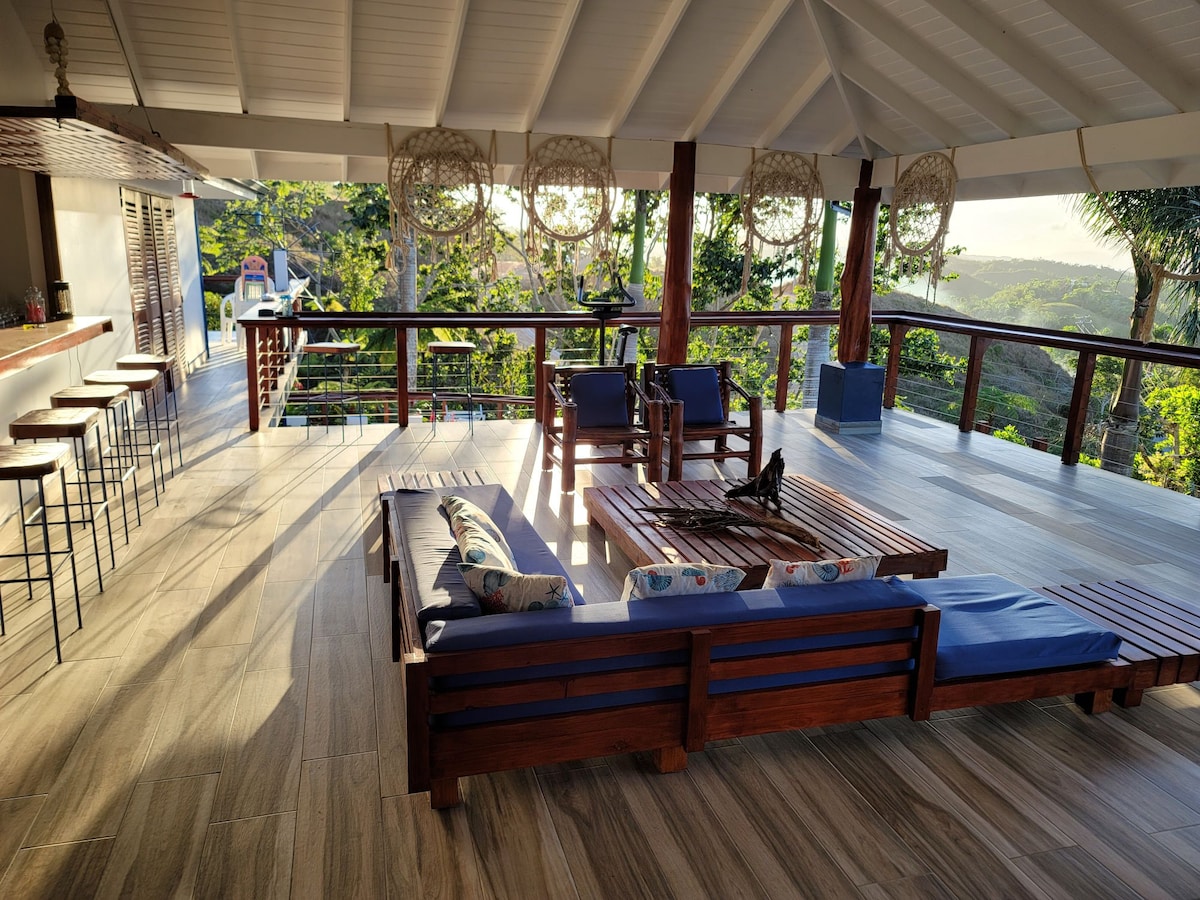 An open-air living space features wooden lounge furniture arranged around a central gathering area. High ceilings and large windows allow natural light to fill the space, offering views of the surrounding greenery. Bar stools line a counter in the background, enhancing the inviting atmosphere.