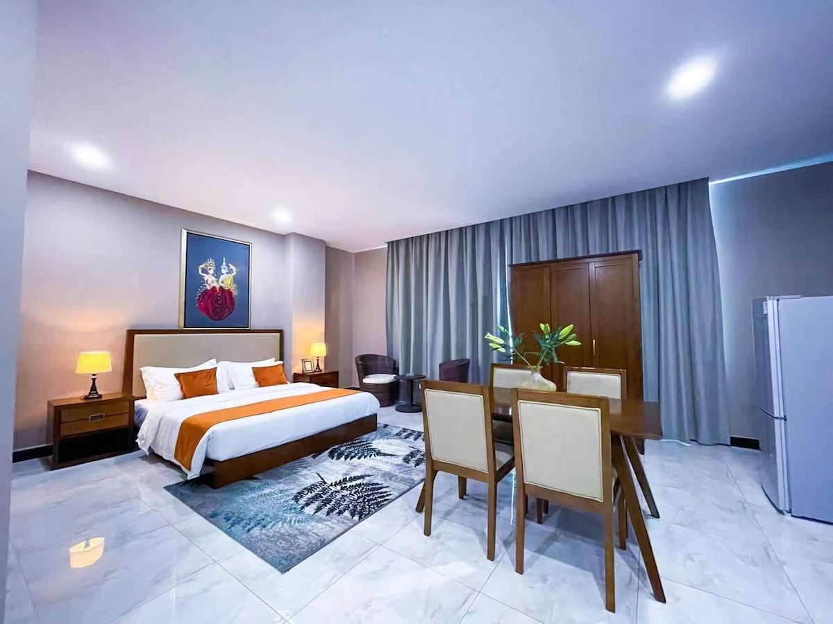 A spacious room features a large bed with crisp white linens and accent pillows. A dining table with four chairs is positioned nearby, beside a pair of cozy armchairs. Soft lighting from lamps enhances the neutral color palette, and a large piece of artwork adds a touch of color.