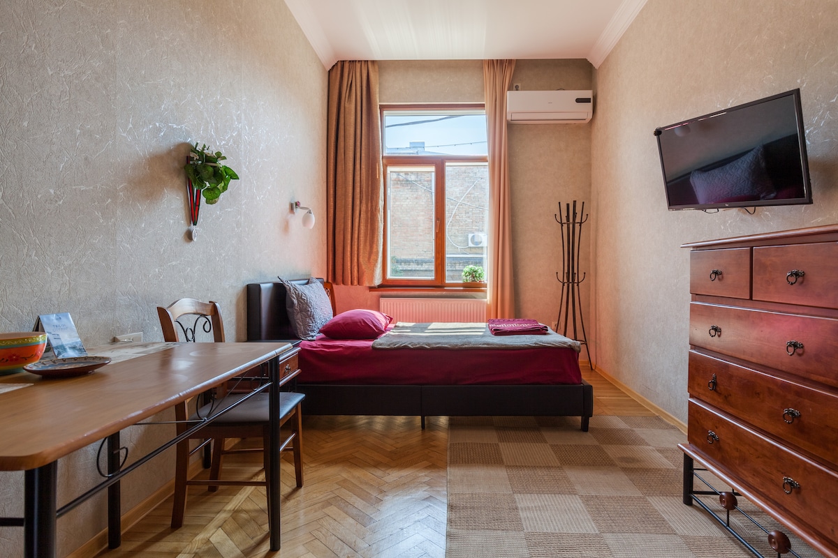 The studio features a single bed with dark bedding against a light wall. A wooden dresser and a small table with chairs are visible. Natural light enters through a large window with light curtains. An air conditioning unit and a television are mounted on the wall.