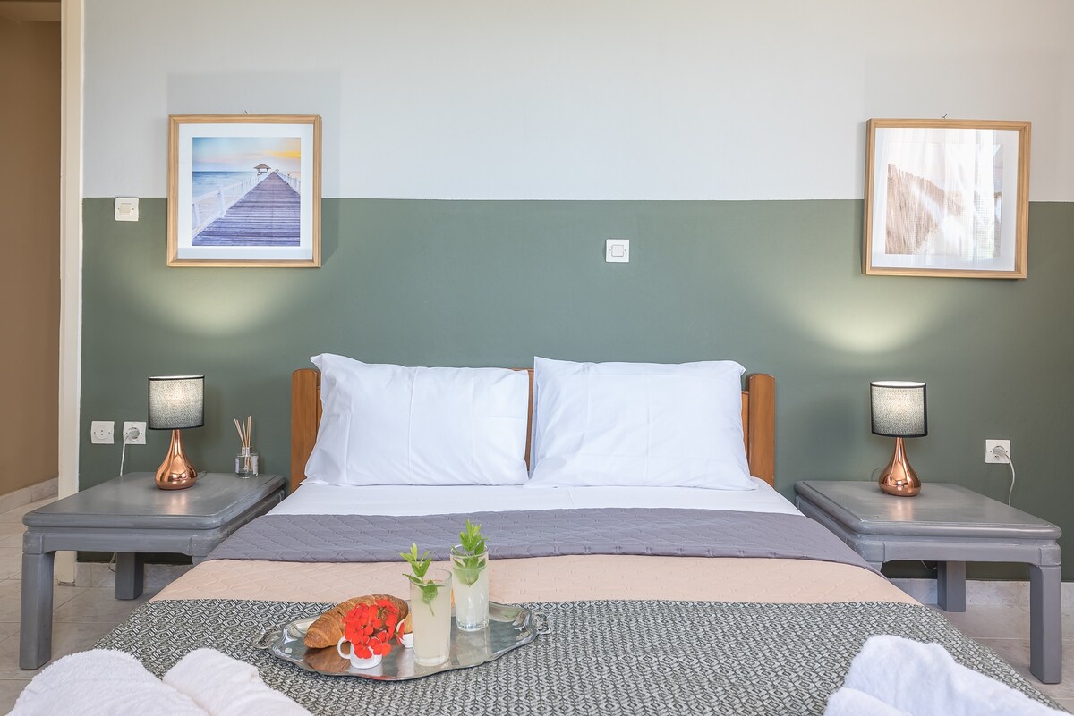 A spacious bedroom is highlighted with two large beds, each flanked by modern bedside tables and lamps. Soft natural light illuminates the room, complemented by green accent walls and framed artwork. A decorative tray with fresh flowers and pastries adds an inviting touch.