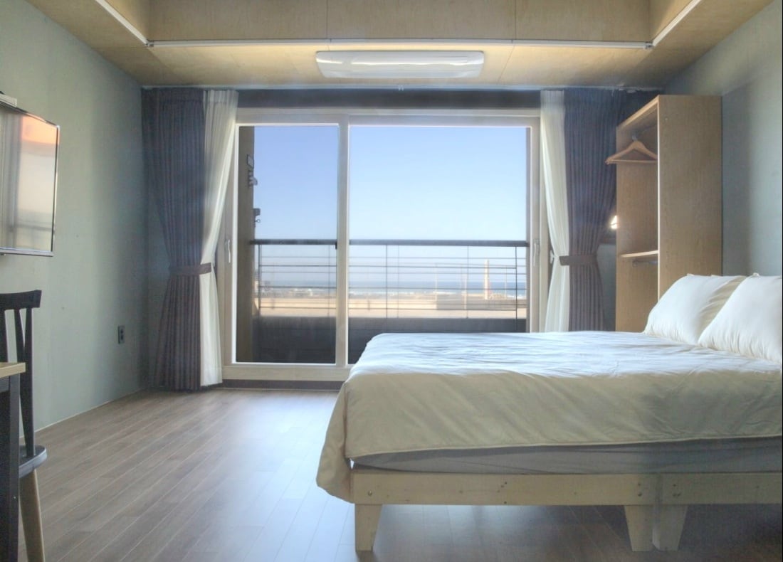 A bright and spacious room features a comfortable bed with crisp white linens. Large glass doors open to a balcony, allowing ample natural light. A minimalistic design is complemented by a television and a wooden shelving unit, enhancing the room's functional layout.
