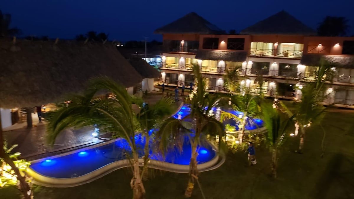 A serene evening scene features a beautifully illuminated pool surrounded by lush palm trees. The resort buildings, adorned with thatched roofs, are visible in the background, enhancing the peaceful ambiance of the outdoor area.
