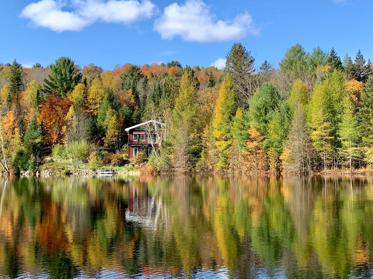 A serene view of a chalet is captured across a lake, surrounded by vibrant autumn foliage. The clear water reflects the colorful trees and the house, creating a harmonious natural scene. Fluffy clouds drift across a bright blue sky, enhancing the tranquil atmosphere.
