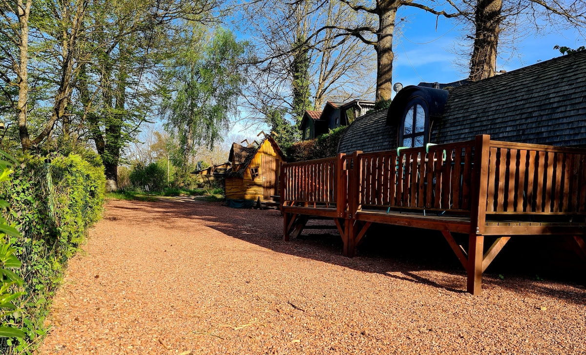 A gravel pathway leads to a uniquely shaped cabin surrounded by trees, with a wooden deck extending towards the front. Nearby, a small wooden structure is partially visible, adding to the natural charm of the setting.