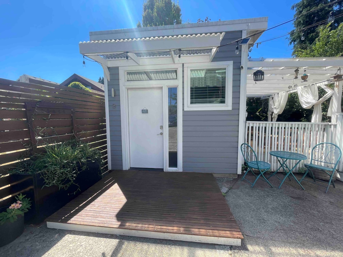 The exterior showcases a welcoming entrance with a modern door and a small wooden deck. Two teal bistro chairs are positioned at a table nearby, surrounded by lush greenery and a fence, providing a sense of privacy.