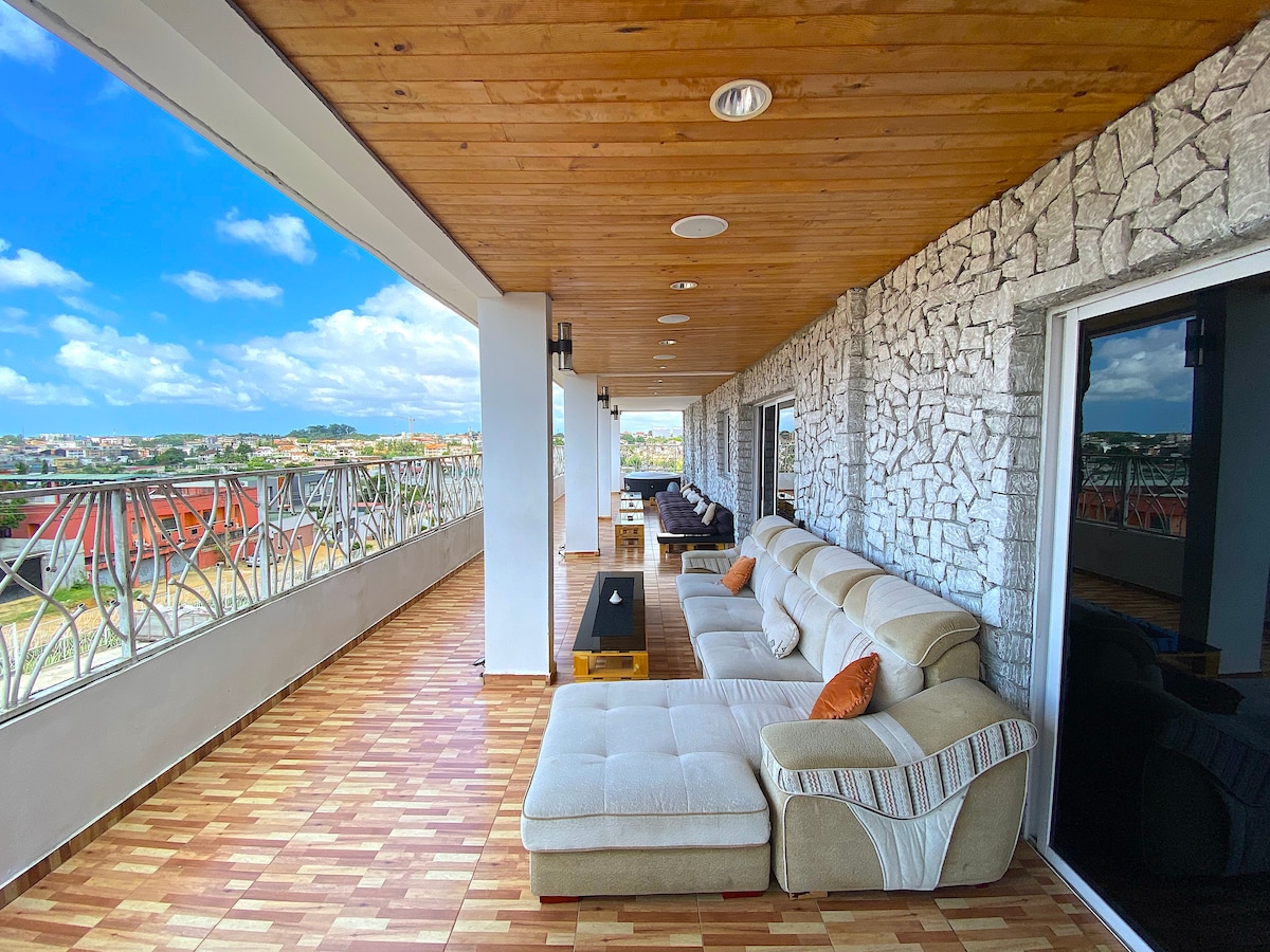 A spacious balcony is shown, featuring a large, L-shaped couch with accent pillows, providing ample seating. Elegant stone walls contrast with the wooden ceiling. Large sliding doors lead to the interior, with a stunning view of the surrounding area visible beyond.