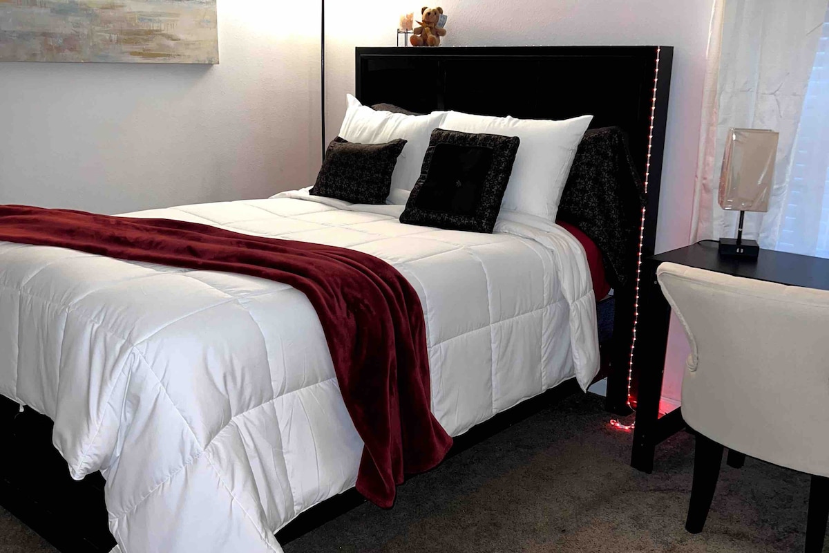 A neatly arranged bedroom is featured with a spacious bed, dressed in white linens and decorative pillows. A soft, burgundy throw is draped across the bed. A bedside table, accompanied by a stylish lamp, complements the modern decor.