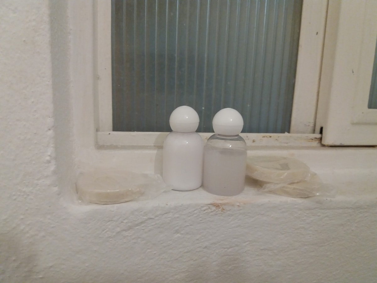 A clean bathroom counter features two modern bottles, one opaque white and the other clear, positioned closely together. Several wrapped soap bars are neatly arranged beside them, all set against a white wall and a frosted glass window providing soft light.