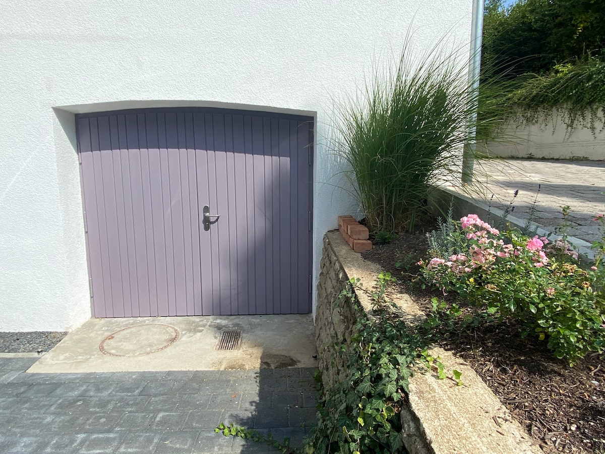 A purple entrance door stands against a white wall, adjacent to a landscaped area filled with pink flowers and greenery. A stone border outlines the flower bed, while a paved pathway leads towards the entrance.