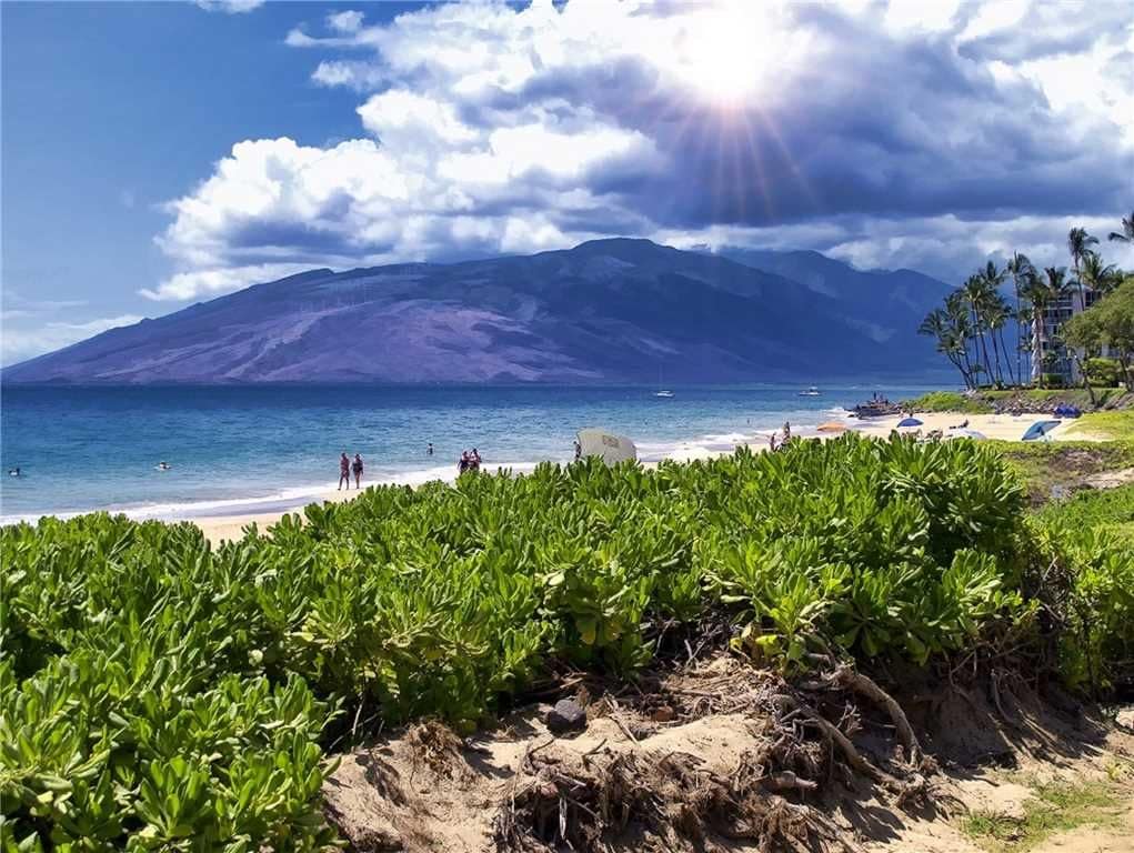Lush green vegetation frames a sandy beach along the coastline. Gentle waves can be seen lapping at the shore while people enjoy the water under a partly cloudy sky. Majestic mountains rise in the background, adding to the serene coastal scenery.