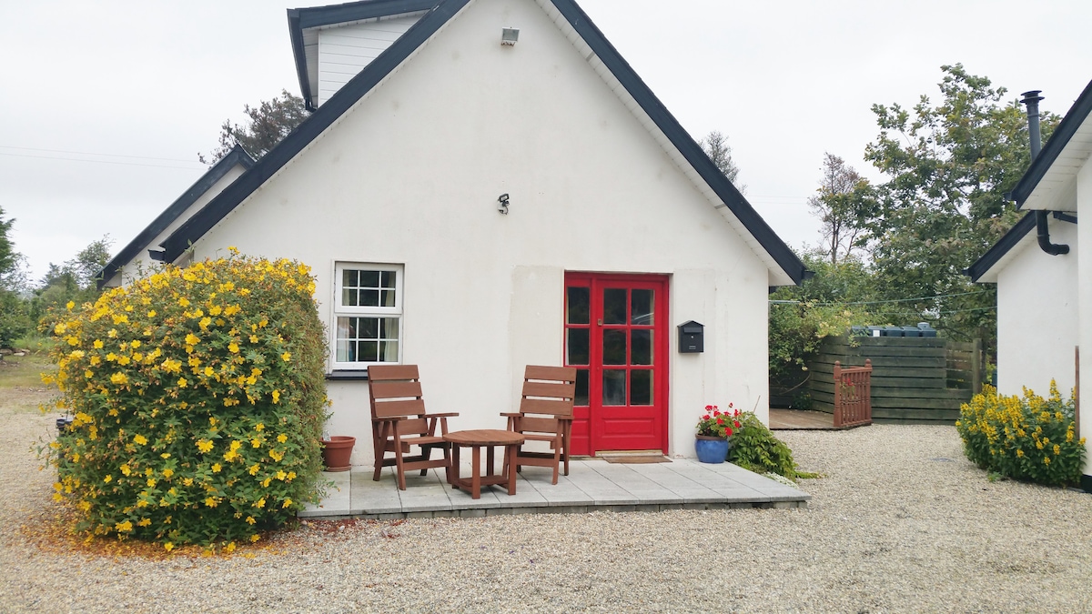 The exterior of a cozy cottage presents a welcoming red door and large windows framed by white walls. Two wooden chairs are positioned on a stone patio, surrounded by flowering shrubs and gravel. A small potted plant sits near the entrance, adding a touch of greenery.