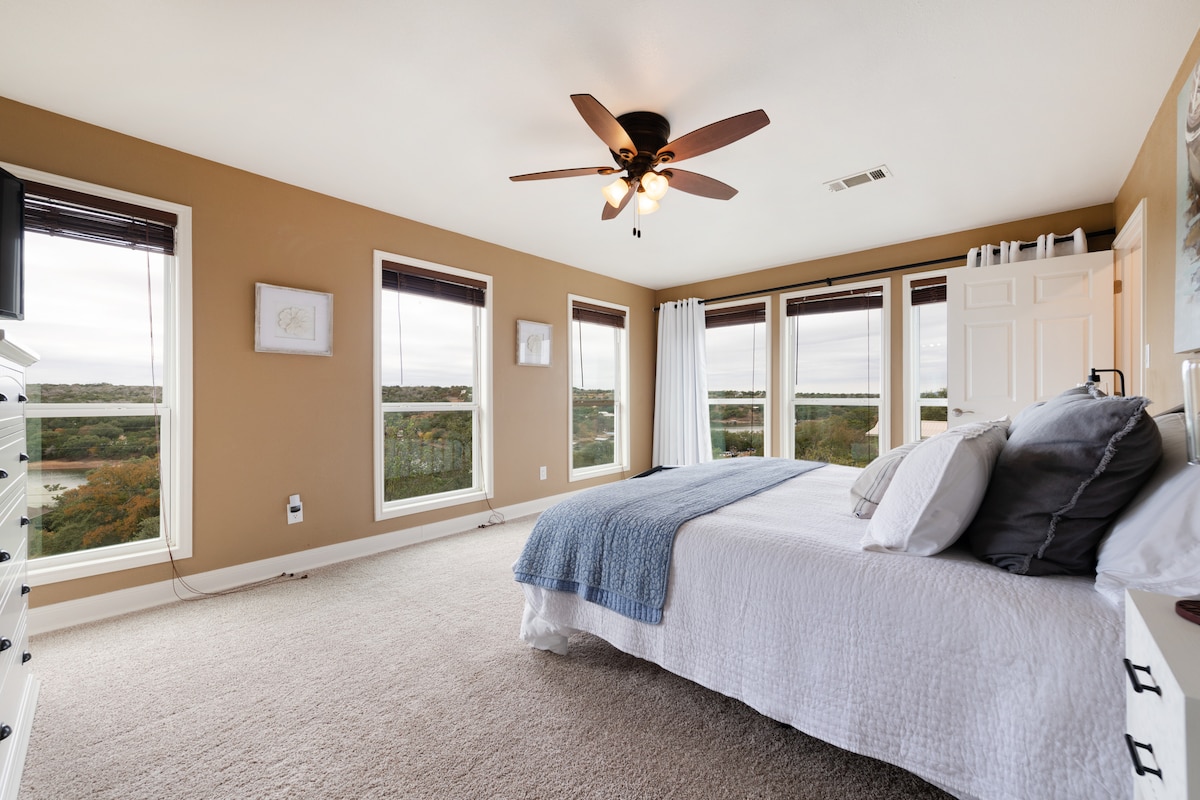 The master bedroom features large windows offering expansive views of the surrounding landscape. A spacious bed is adorned with soft linens, and a ceiling fan hangs above. Neutral walls and carpet create a calming environment, enhanced by natural light flowing in through the windows.