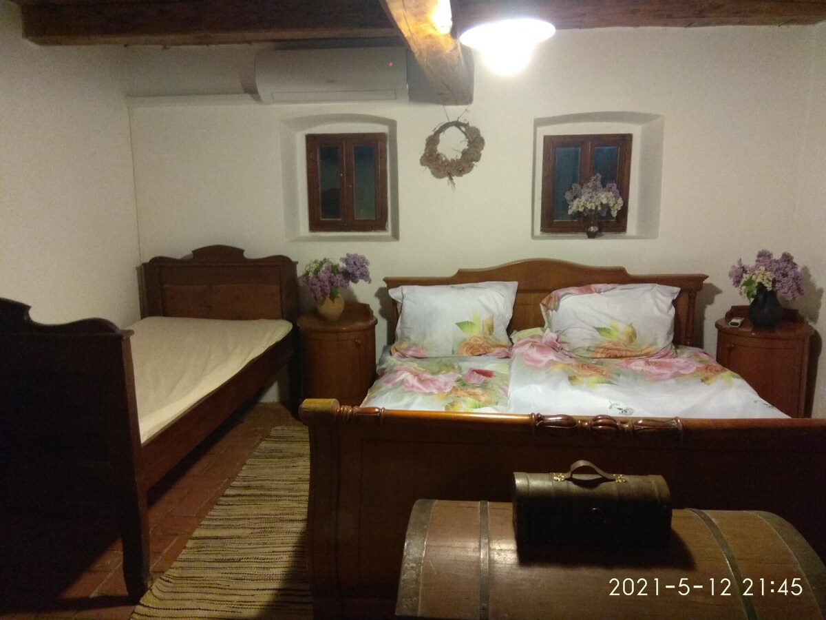 A cozy bedroom features a spacious double bed with floral bedding and a single bed nearby. Wooden furniture adds a rustic charm, with a trunk at the foot of the beds. Natural light enters through two small windows, enhancing the serene ambiance.