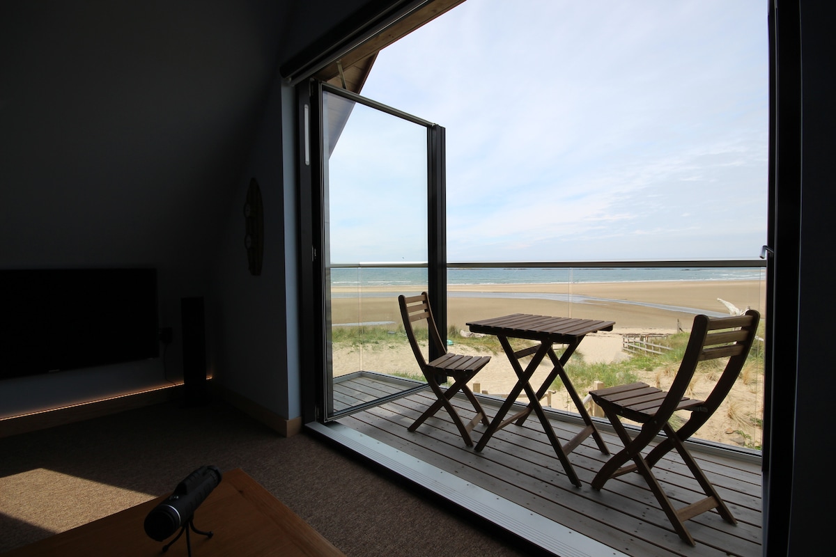 A private balcony is visible, featuring a small wooden table and two chairs, offering unobstructed views of the beach and ocean. Large patio doors open to enhance the connection with the seaside. The natural light gently illuminates the space.