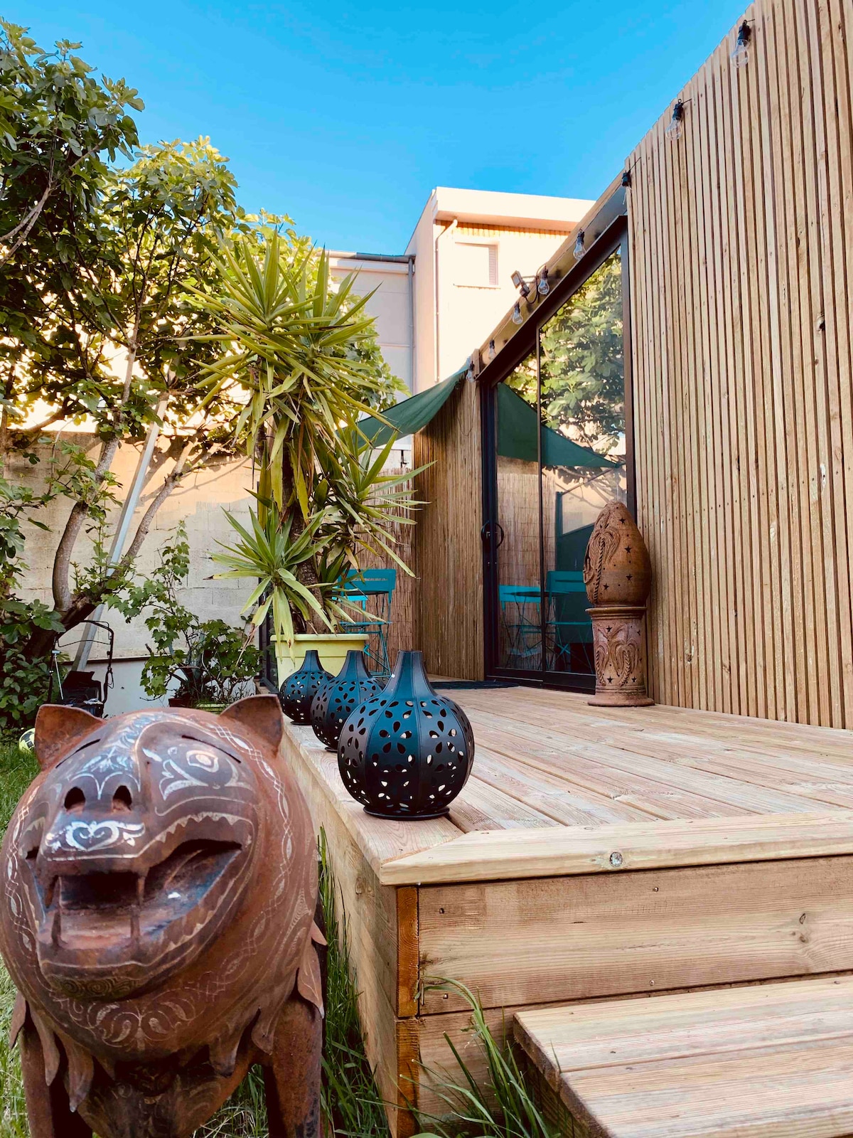 A wooden deck is featured in the foreground, surrounded by greenery. Decorative items, including carved sculptures and vases, add character to the space. The sliding glass door offers a view into the interior, while natural sunlight enhances the inviting atmosphere.