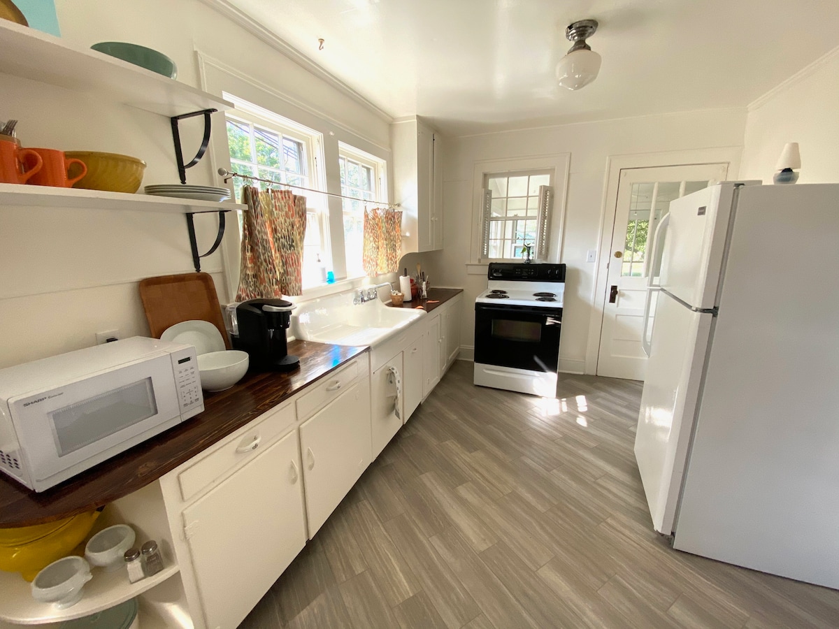The well-equipped kitchen features white cabinetry and a stainless steel refrigerator. A microwave and coffee maker are positioned on a countertop. Natural light streams through multiple windows, illuminating the space with a light, airy feel. The flooring is a neutral, stylish design.