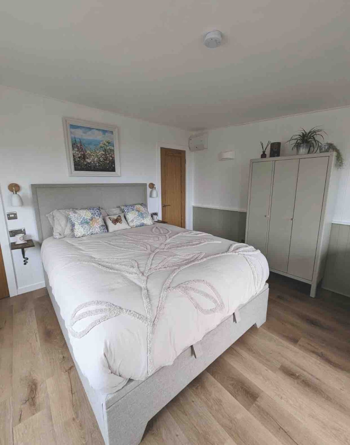 A bright bedroom is furnished with a spacious bed dressed in a soft, light-colored coverlet. Decorative pillows add a touch of color. A wardrobe stands nearby, and artwork is displayed on the wall, enhancing the room's inviting atmosphere.