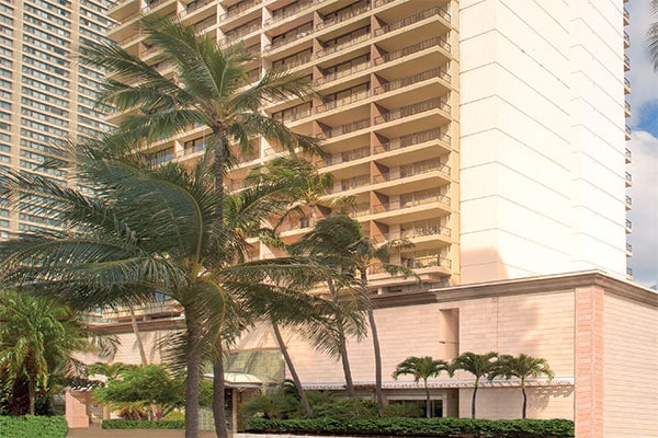 The resort building is shown, featuring multiple floors with balconies. Lush palm trees surround the entrance and landscaping, creating a tropical atmosphere. The structure's light-colored facade contrasts with the greenery, and large windows are visible on each level.
