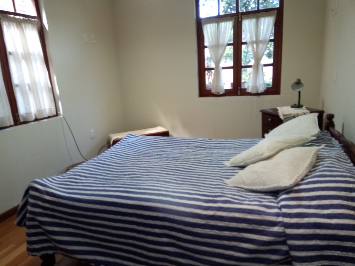 A cozy bedroom features a spacious bed adorned with striped blankets and soft pillows. Two windows allow natural light to fill the room, with sheer curtains softly filtering the view outside. A small bedside table and a wooden stool add functionality to the space.