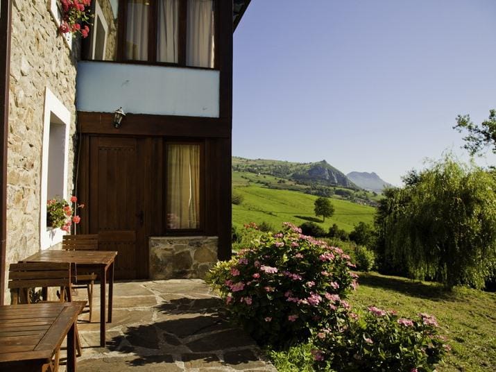 The exterior of the traditional stone house is framed by vibrant flowers and greenery, with rustic wooden tables positioned on a stone patio. The landscape features rolling hills and distant mountains under a clear blue sky.