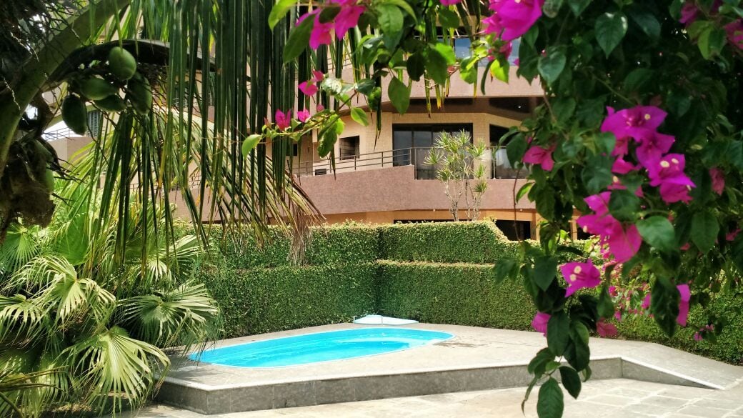 A private swimming pool is surrounded by lush greenery and blooming pink flowers. The pool area is framed by well-maintained hedges, providing a sense of seclusion. The backdrop features a multi-story building with balconies, adding to the serene ambiance of the outdoor space.