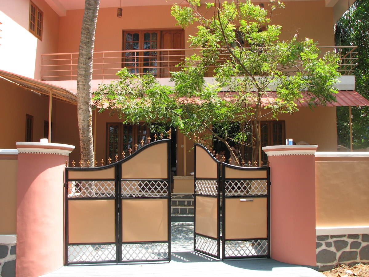 A welcoming entrance features an open gate leading to a spacious property. The facade is painted in warm tones, highlighting large windows and a balcony above. Lush greenery surrounds the area, providing a pleasant natural setting.