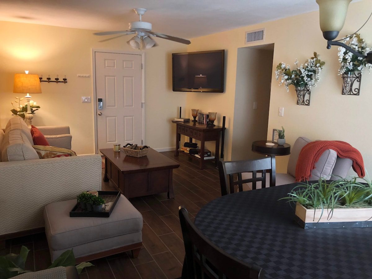 The living and dining area features a neutral-toned sofa and armchair, paired with a dark wood coffee table. A flat-screen TV is mounted on the wall, while decorative accents include plants and wall-mounted flower arrangements. A round dining table with chairs is also present.