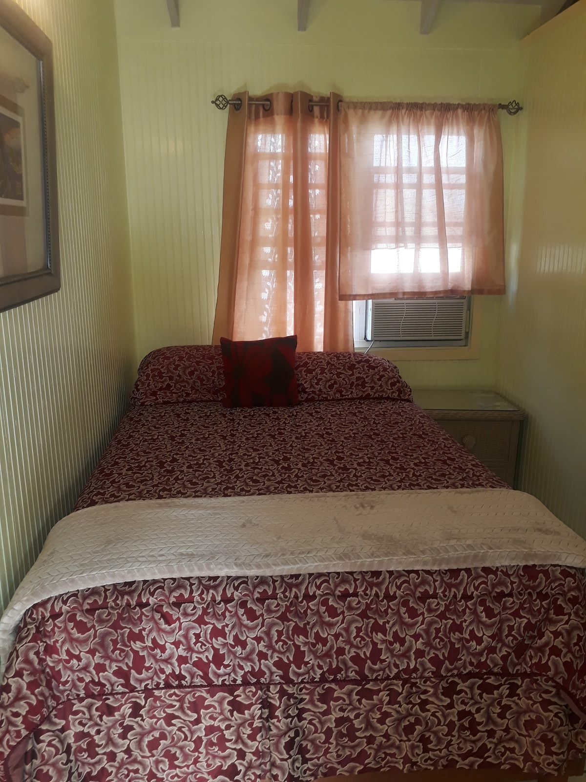 A compact sleeping area features a twin bed with a patterned bedspread and a decorative pillow. A small nightstand is positioned alongside the bed, with an air conditioning unit mounted in the window. Soft light filters through sheer curtains, creating a warm ambiance.