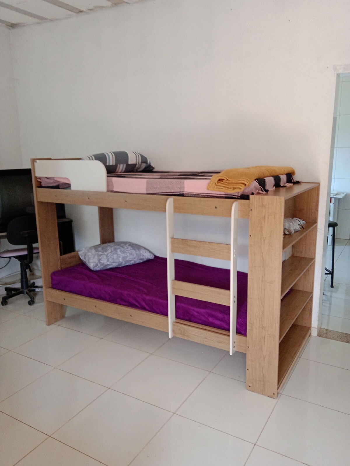 A wooden bunk bed is featured, showcasing a lower sleeping area with a purple blanket and a gray pillow. The upper level is decorated with striped and solid-colored bedding. A simple wooden shelving unit adds storage space on one side.