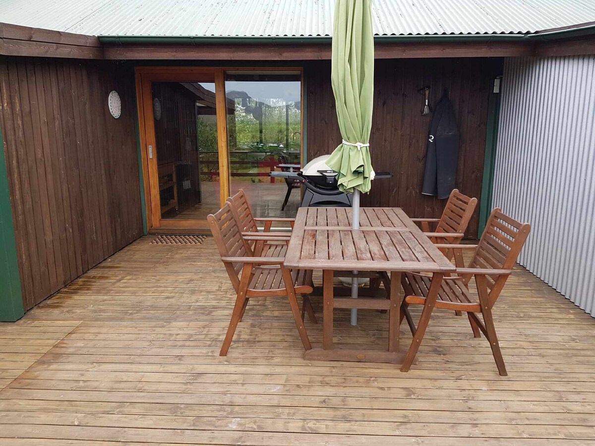 A wooden deck is presented with a rectangular table surrounded by six matching chairs. A green umbrella stands at the center, providing shade. The adjoining structure features large glass doors that lead inside, enhancing the connection between indoor and outdoor spaces.