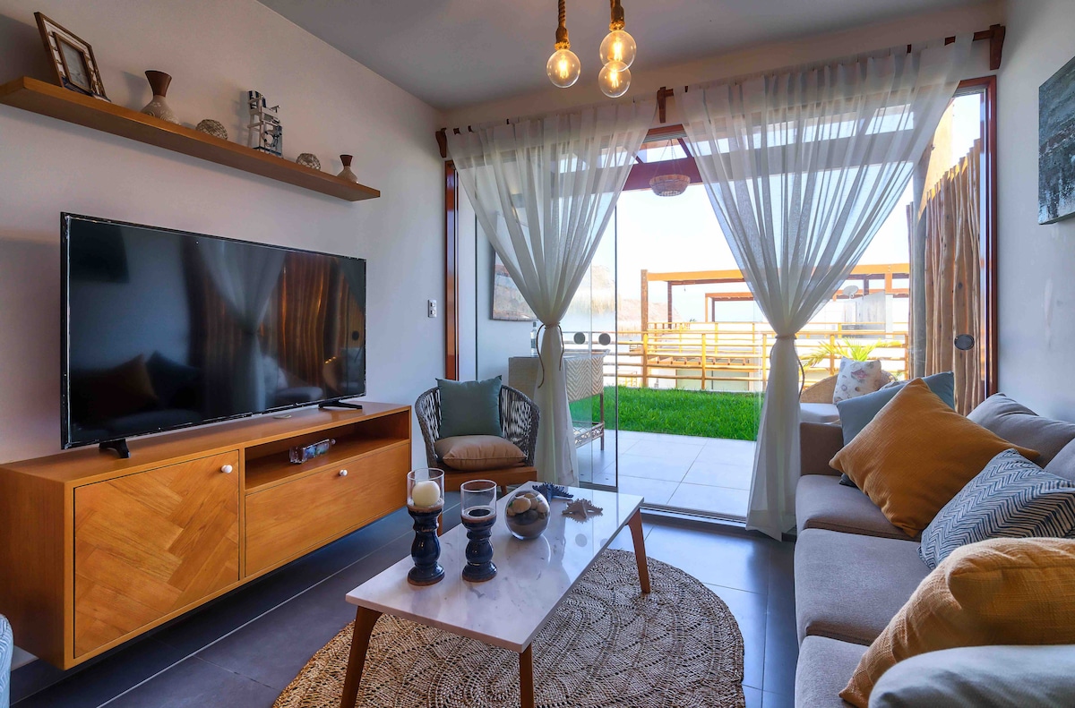 A living area is shown featuring a comfortable sofa adorned with pillows and a coffee table. A large television stands on a wooden console, while light-colored curtains frame the view of a grassy outdoor space through sliding glass doors.