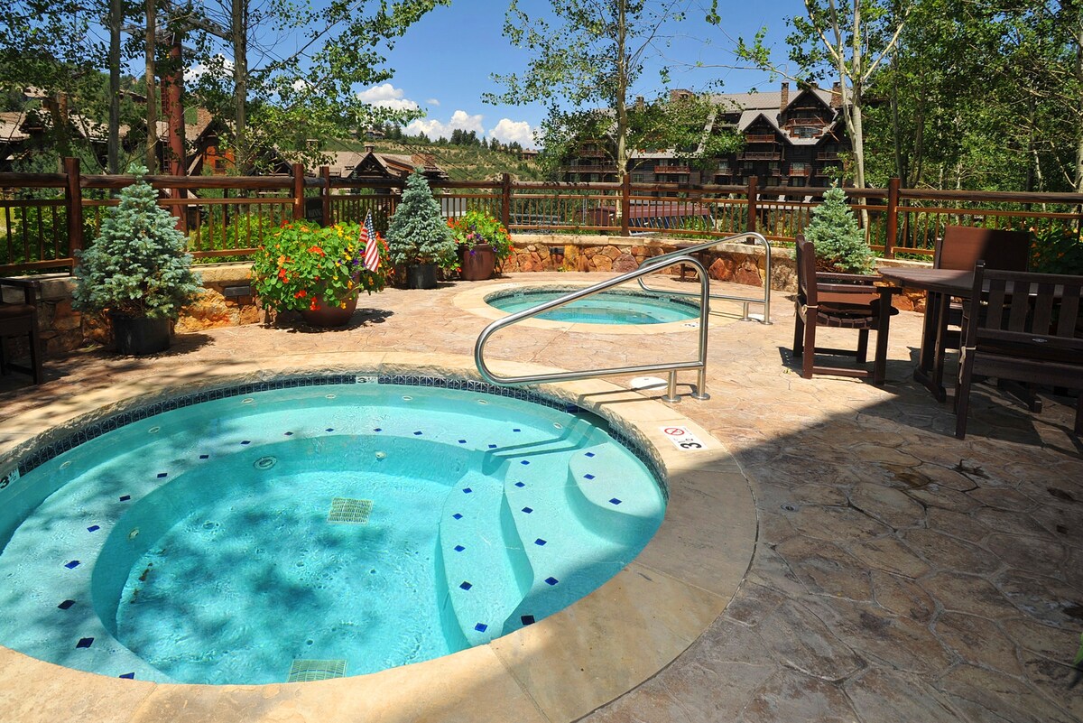 Two inviting hot tubs are situated outdoors, surrounded by natural stone. Lush greenery, including small pine trees and colorful flower pots, enhance the space. A wooden railing provides a warm boundary, and hillside structures can be glimpsed in the background under a clear blue sky.