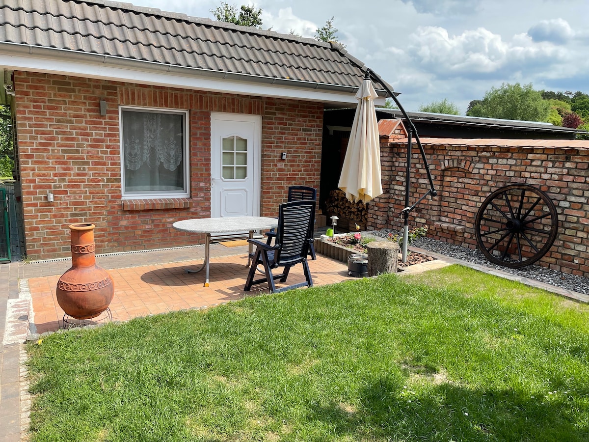 An outdoor patio features a round table surrounded by two black chairs. A large umbrella provides shade, while a clay pot and a rustic wooden stump add character. A stone wall and an antique wheel enhance the garden's charm, set in a grassy area.