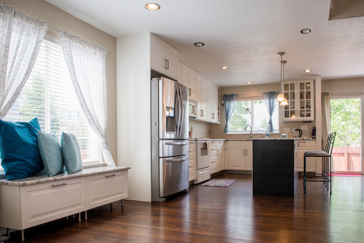 A spacious kitchen area is presented, featuring modern appliances and ample cabinetry. Sunlight filters through the large windows, illuminating a comfortable seating area with pillows. A dark island with bar seating adds functionality to the inviting space, complemented by light-colored countertops and wood flooring.