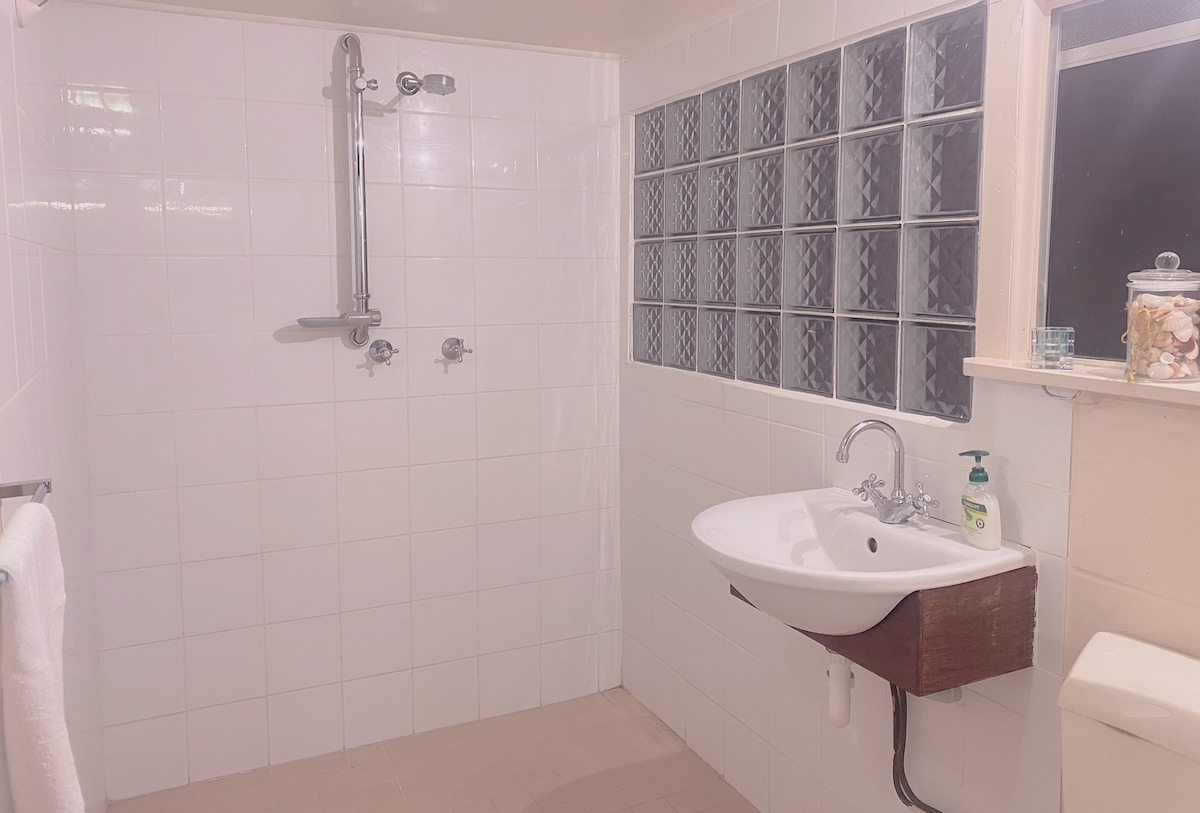 A clean bathroom is presented with white tiled walls and a large walk-in shower equipped with a handheld showerhead. A modern sink with a wooden shelf is featured alongside a tall window displaying decorative glass blocks. A towel is neatly hung on the side.