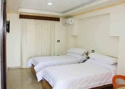 A well-lit room features two spacious beds with white bedding, positioned against a light-colored wall. Natural light filters through sheer curtains, and an air-conditioning unit is mounted above. A comfortable chair is placed near the entrance.