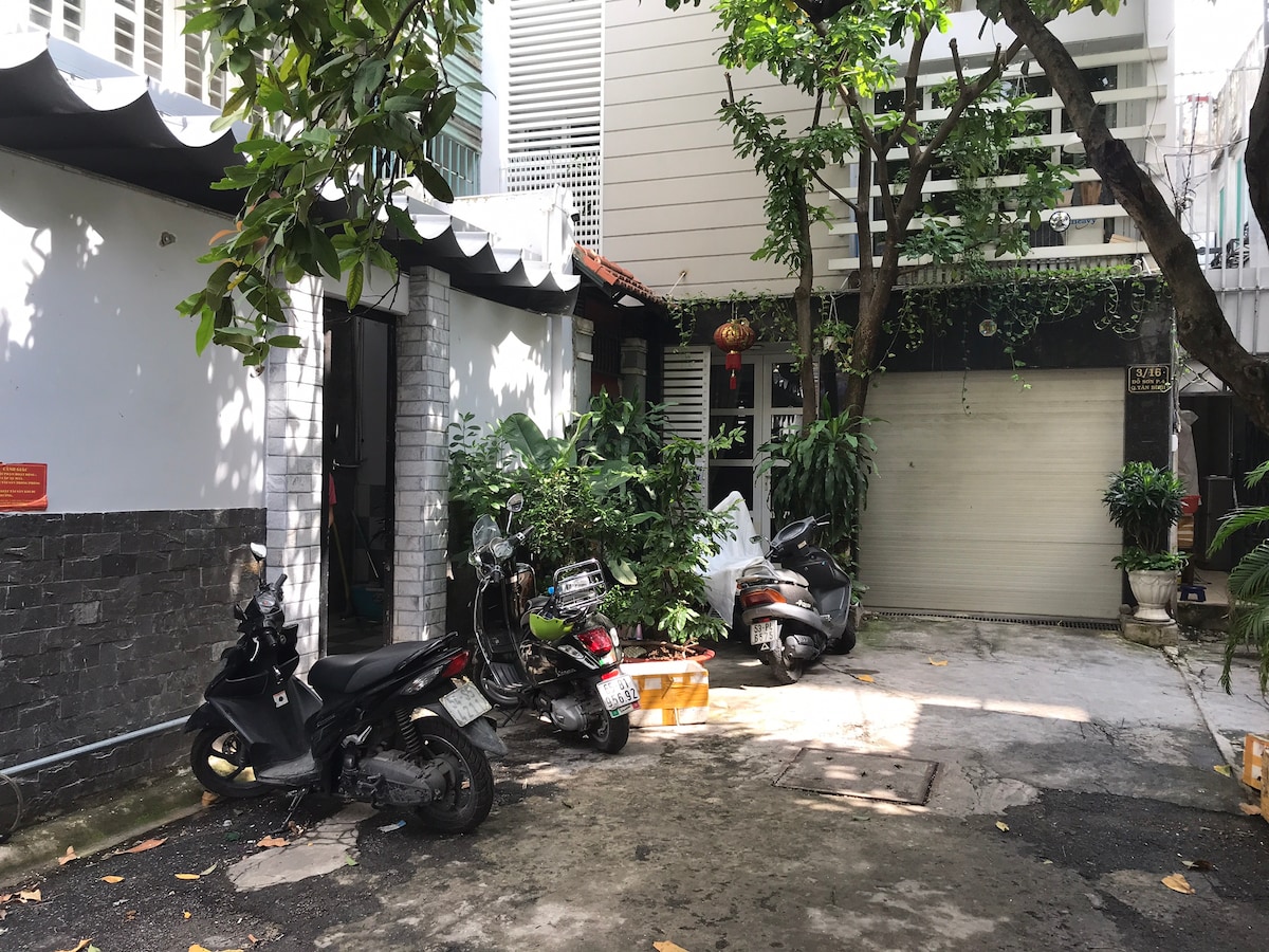 A peaceful outdoor area is visible, featuring a small parking space for motorcycles. Lush green plants are scattered throughout, providing a natural touch. The surroundings include a partially covered entrance, with sunlight filtering through the trees, creating a serene atmosphere.