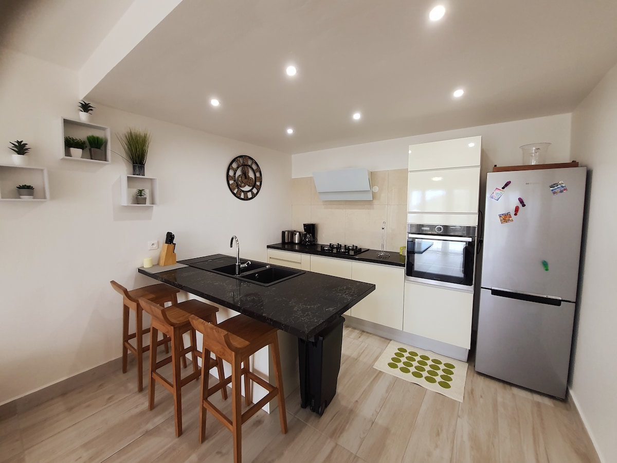 A modern kitchen features sleek cabinetry and a dark countertop. Three wooden stools are arranged at a bar-height area. Appliances include a refrigerator, an oven, and a stovetop. Soft lighting is provided by recessed ceiling fixtures, enhancing the bright and open space.