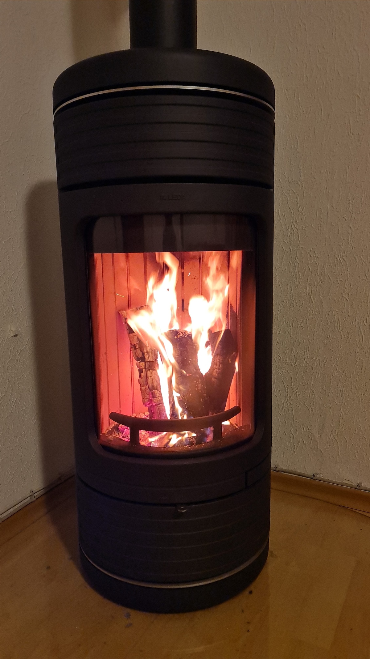 A modern wood-burning stove stands against a textured wall, featuring a cylindrical design. Flames flicker inside, casting warm light against the natural wood flooring surrounding the stove, creating a cozy focal point in the living space.