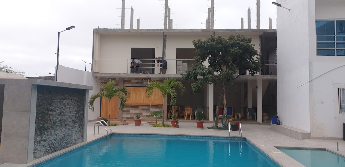 A swimming pool is situated at the forefront, surrounded by tall leafy plants in pots. In the background, a multi-story building is partially constructed, featuring a mix of modern architectural elements. The area is designed for relaxation, with comfortable seating and a tranquil water feature.