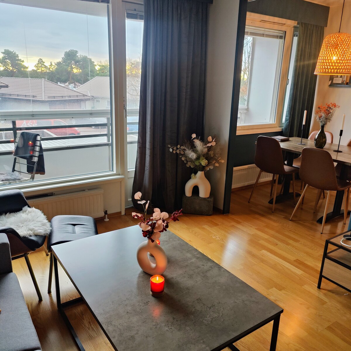 A modern living area features a sleek coffee table with a decorative vase and a candle. Soft seating is arranged near a large window that lets in natural light, while a dining table with chairs is visible in the background.
